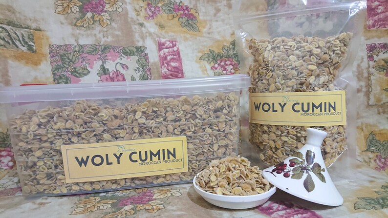 Op de afbeelding: Twee containers met gedroogde, lichtbruine, hele komijnzaadjes. De containers zijn gelabeld met "Woly Cumin Moroccan Product".
