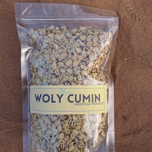 Op de afbeelding: Een doorzichtige plastic zak gevuld met gedroogde, lichtbruine en groene zaden. De zak heeft een geel label met de tekst "WOLY CUMIN MOROCCAN PRODUCT".