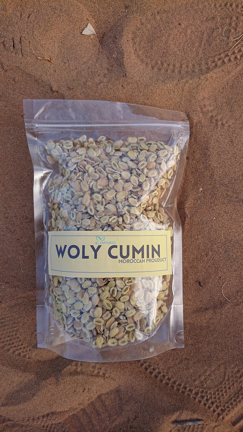 Op de afbeelding: Een doorzichtige plastic zak gevuld met gedroogde, lichtbruine en groene zaden. De zak heeft een geel label met de tekst "WOLY CUMIN MOROCCAN PRODUCT".