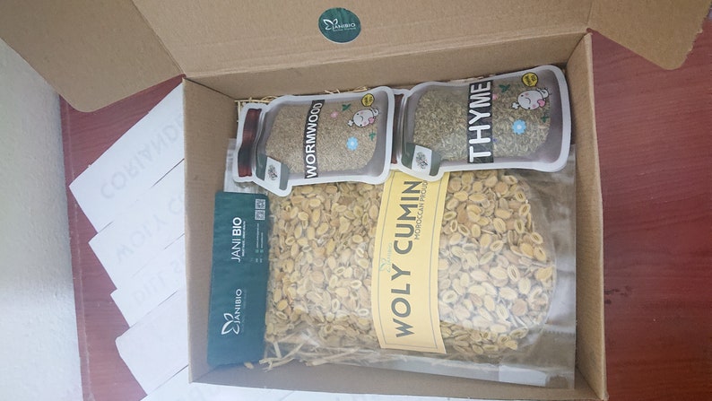 Op de afbeelding: Een kartonnen doos gevuld met verschillende gedroogde kruiden en specerijen. De doos bevat een zak "Holy Cumin" met een geel etiket, een zak "Wormwood" met een wit etiket en een zak "Thyme" met een wit etiket. De doos is gevuld met bruin verpakkingsmateriaal.