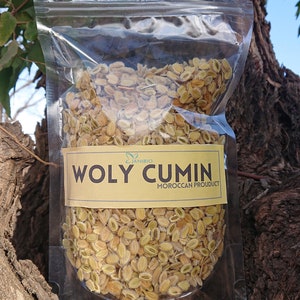 Op de afbeelding: Een doorzichtige plastic zak gevuld met gedroogde, lichtbruine zaden met het label "WOLY CUMIN MOROCCAN PRODUCT".
