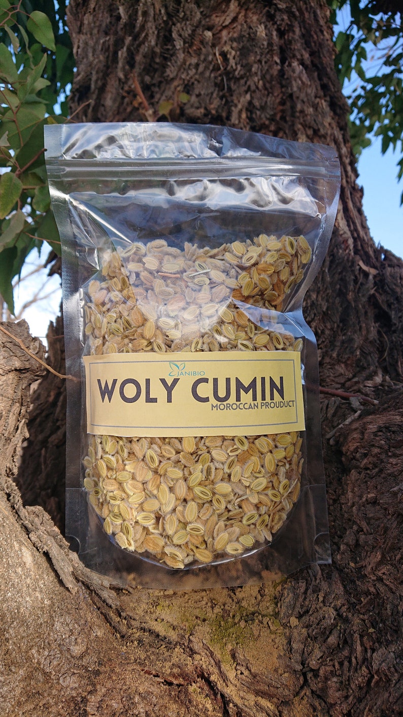 Op de afbeelding: Een doorzichtige plastic zak gevuld met gedroogde, lichtbruine zaden met het label "WOLY CUMIN MOROCCAN PRODUCT".