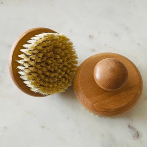 Könnte beinhalten: Zwei Körperbürsten aus Holz mit Naturborsten. Eine Bürste hat einen runden Griff und die andere einen Knopfgriff.