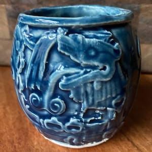May include: A small, round, blue ceramic pot with a raised design of stylized dragons and swirls. The pot has a white rim and a glossy finish.