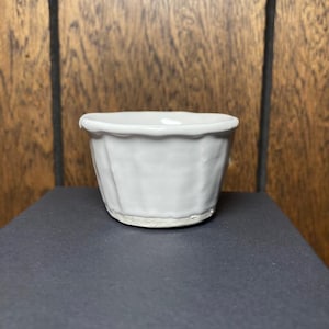May include: A small, white ceramic bowl with a textured exterior and a slightly ruffled rim. The bowl is cylindrical in shape and sits on a dark surface, with a wooden background.