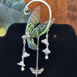 May include: A silver ear cuff with a fairy wing design. The wings are green and brown with glitter accents. Dangling from the wings are chains with small white flower and pearl charms, and a moth charm. A crescent moon with a star is at the top.