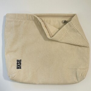 Pode incluir: Bolsa tote de veludo cotel&ecirc; creme com linhas verticais. A bolsa tem uma parte superior dobrada com um fecho de metal e a palavra "BXBIE" impressa verticalmente em preto na lateral.