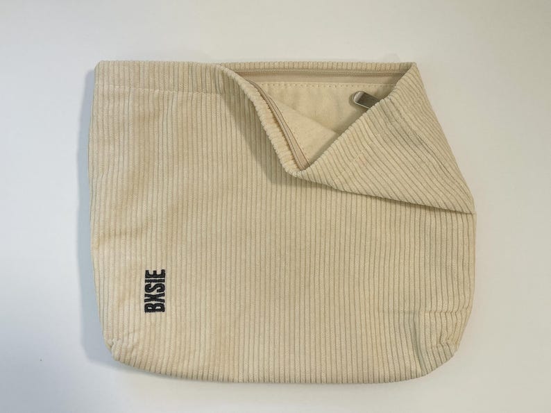 Pode incluir: Bolsa tote de veludo cotel&ecirc; creme com linhas verticais. A bolsa tem uma parte superior dobrada com um fecho de metal e a palavra "BXBIE" impressa verticalmente em preto na lateral.