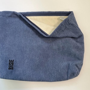 Pode incluir: Uma bolsa tote azul de veludo cotel&ecirc; com formato retangular. A bolsa tem fecho de z&iacute;per e interior de cor clara. A palavra "BXSIE" est&aacute; impressa verticalmente em preto no lado inferior esquerdo.