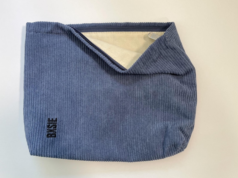 Pode incluir: Uma bolsa tote azul de veludo cotel&ecirc; com formato retangular. A bolsa tem fecho de z&iacute;per e interior de cor clara. A palavra "BXSIE" est&aacute; impressa verticalmente em preto no lado inferior esquerdo.