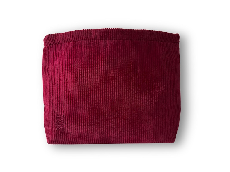 Pode incluir: Uma bolsa de veludo cotel&ecirc; bord&ocirc; com formato retangular. A bolsa tem uma textura canelada e um fecho com cord&atilde;o. O canto inferior esquerdo apresenta um logotipo bordado. O fundo &eacute; branco.