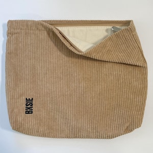 Pode incluir: Bolsa de veludo cotel&ecirc; bege com a parte superior dobrada, revelando um interior creme. A bolsa tem nervuras verticais e a palavra "Bysie" impressa verticalmente em preto no lado inferior esquerdo. Um z&iacute;per de metal prateado &eacute; vis&iacute;vel.