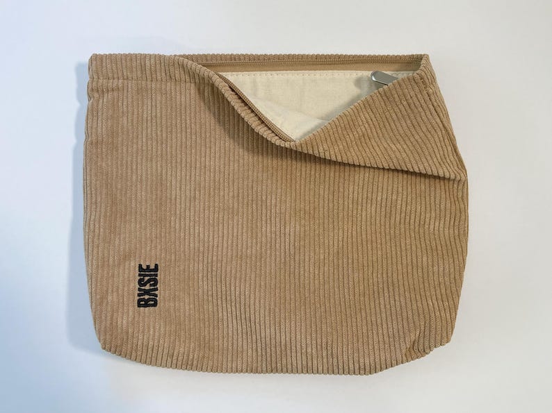 Pode incluir: Bolsa de veludo cotel&ecirc; bege com a parte superior dobrada, revelando um interior creme. A bolsa tem nervuras verticais e a palavra "Bysie" impressa verticalmente em preto no lado inferior esquerdo. Um z&iacute;per de metal prateado &eacute; vis&iacute;vel.