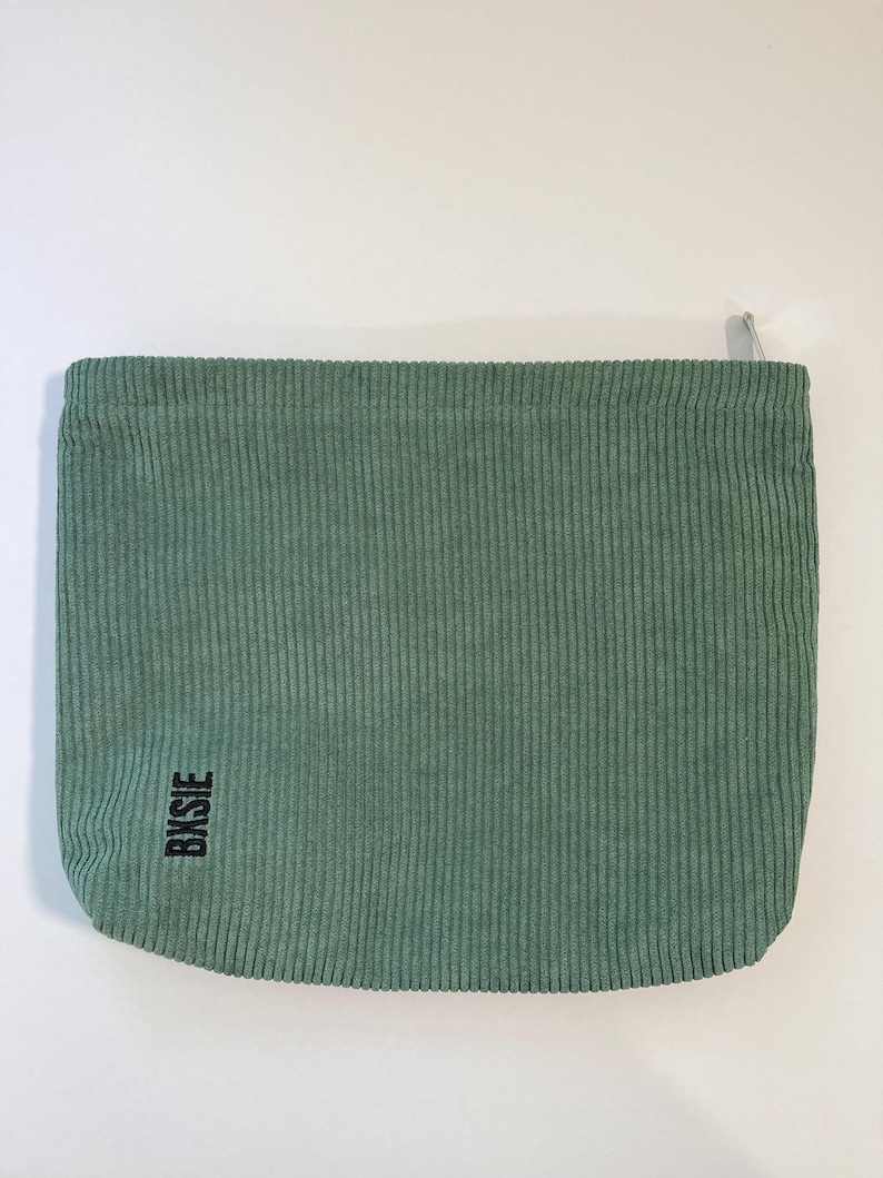 Pode incluir: Uma bolsa de veludo cotel&ecirc; verde s&aacute;lvia com fecho de correr. A bolsa tem uma textura canelada vertical e a palavra "BKSJE" impressa em preto no canto inferior esquerdo. Fundo branco liso.