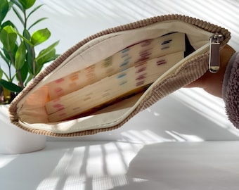 Pode incluir: Uma bolsa com zíper de veludo cotelê bege, aberta para revelar vários leques de papel coloridos. O forro interno é de tecido bege claro. O puxador do zíper é metálico.