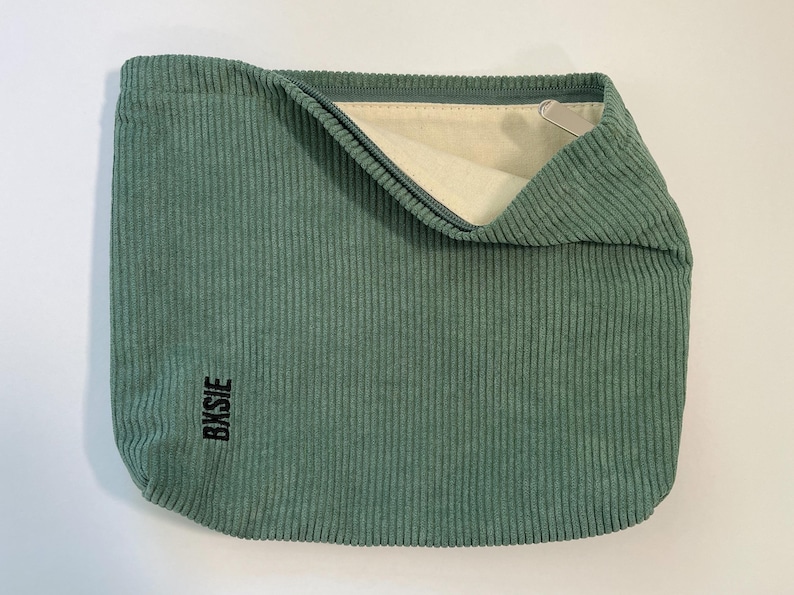 Pode incluir: Uma bolsa de veludo cotel&ecirc; verde s&aacute;lvia com fecho de correr. A bolsa tem formato retangular e aspecto macio e texturizado. A palavra "Bksie" est&aacute; impressa em preto no canto inferior esquerdo. O interior &eacute; bege claro.