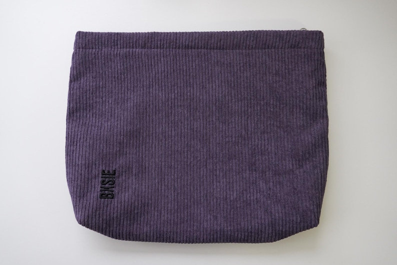 Pode incluir: Uma bolsa retangular de veludo cotel&ecirc; roxo com nervuras verticais. A bolsa tem um fecho de correr na parte superior e a palavra "BKSJE" &eacute; bordada em preto no canto inferior esquerdo. A bolsa est&aacute; sobre um fundo branco.