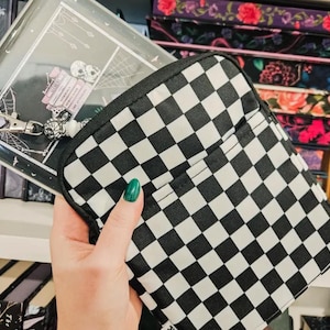 May include: A black and white checkered pouch with a black zipper and trim. The pouch is held in front of a notebook with a skull and spiderweb design. Other notebooks with floral patterns are in the background.