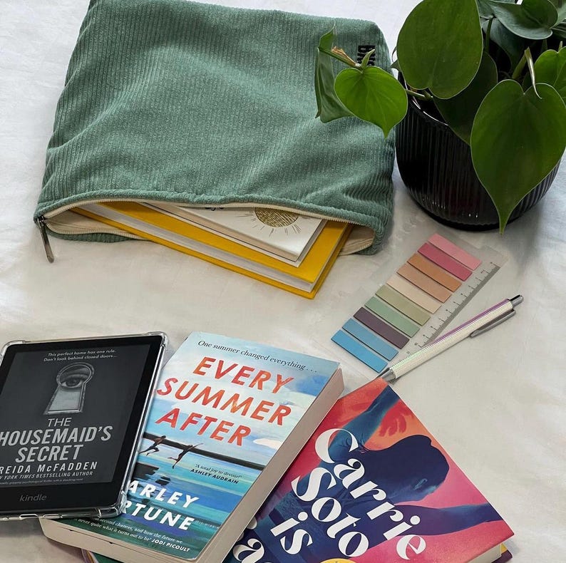 May include: A collection of books, a Kindle, and stationery are arranged on a white surface. A green corduroy pouch is open, revealing yellow and white books. A potted plant, a pen, and sticky notes complete the scene, suggesting a reading or study theme.