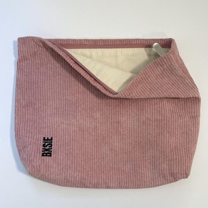 Pode incluir: Uma bolsa de veludo cotel&ecirc; rosa empoeirado com fecho de correr. O interior &eacute; de tecido bege claro. A palavra "Bxsie" est&aacute; impressa verticalmente em preto no lado inferior esquerdo. Um puxador de z&iacute;per de metal prateado &eacute; vis&iacute;vel.