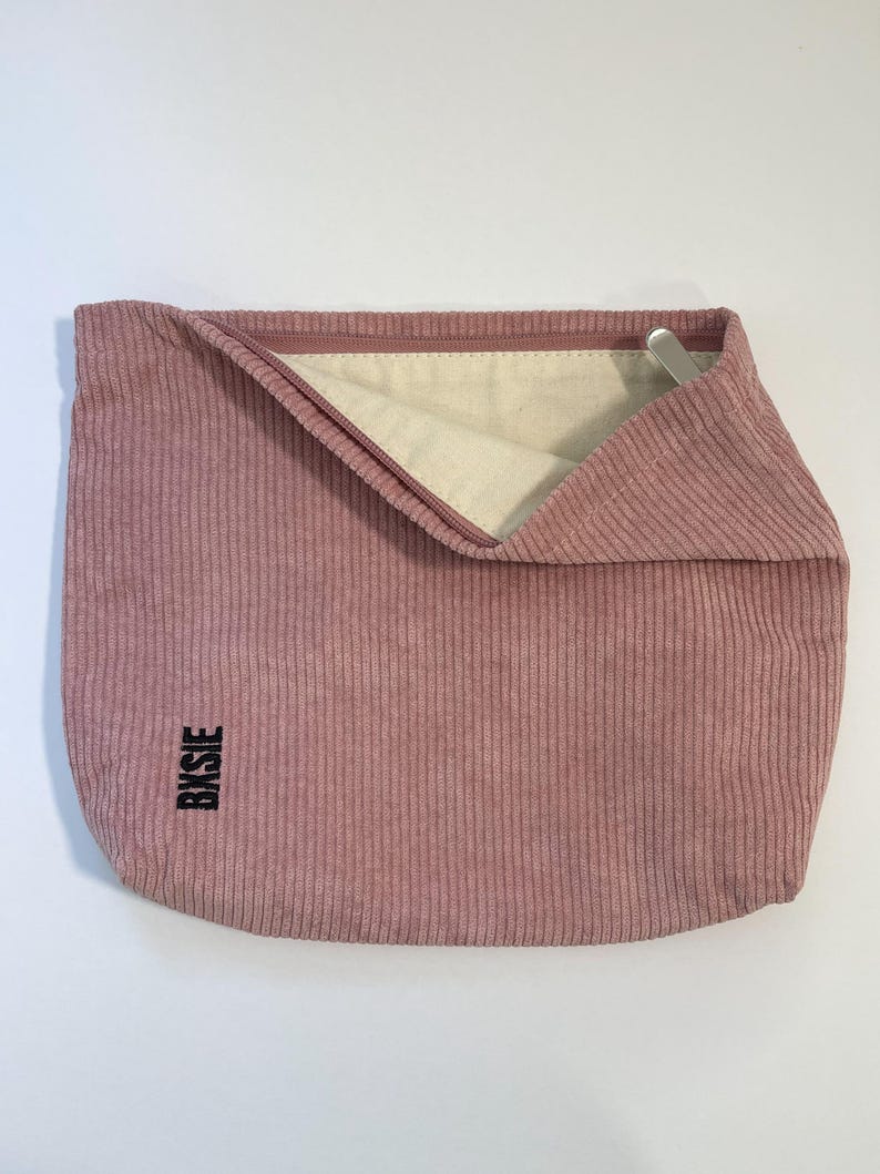 Pode incluir: Uma bolsa de veludo cotel&ecirc; rosa empoeirado com fecho de correr. O interior &eacute; de tecido bege claro. A palavra "Bxsie" est&aacute; impressa verticalmente em preto no lado inferior esquerdo. Um puxador de z&iacute;per de metal prateado &eacute; vis&iacute;vel.