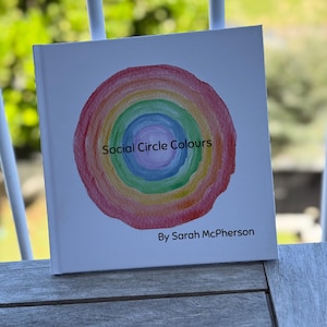 以下が含まれることがあります： サラ・マクファーソン著の「Social Circle Colours」というタイトルのハードカバー本。表紙には、虹色の同心円の水彩デザインが描かれています。本は木製の表面に置かれています。