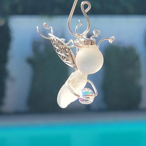 May include: A silver wire angel ornament with a white sea glass body and a clear crystal in its hand. The ornament has a silver wire loop for hanging.