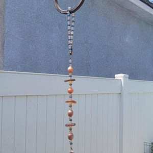 May include: A decorative wind chime with a curved metal hook. The chime features a beaded strand with blue, orange, and brown beads, and a small, patterned bell at the bottom. The background includes a white fence and a blue wall.