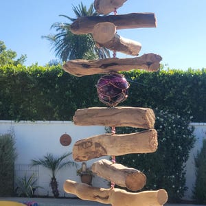 May include: A handcrafted driftwood wind chime with a purple glass orb and pink bead accents. The natural wood pieces are arranged in a spiral design, suspended from a string. The wind chime is displayed outdoors.