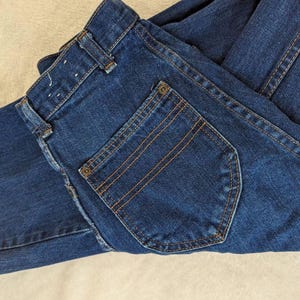 May include: Dark blue denim jeans with a classic five-pocket design. The jeans feature a button and zipper closure, belt loops, and a back pocket with orange stitching. The denim is folded, showing the details of the stitching and construction.