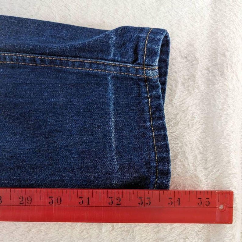 May include: Close-up of a section of dark blue denim jeans. The jeans have a folded hem with visible yellow stitching. A red ruler is placed along the bottom, showing measurements from 28 to 35 inches.