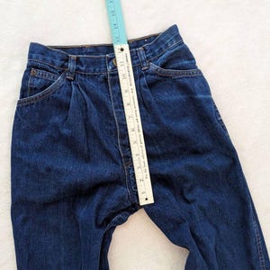 May include: Dark blue denim jeans with a ruler measuring the inseam. The jeans have a classic five-pocket design, a button and zipper closure, and pleats at the waist. The ruler shows a measurement of approximately 11 inches.