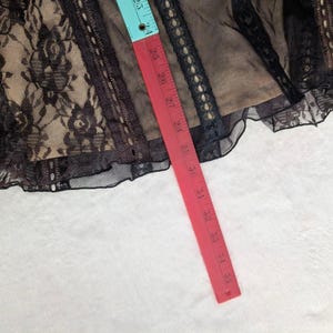 Puede incluir: Tela de encaje negro con una capa base beige, con patrones florales y adornos decorativos. Una cinta m&eacute;trica roja y azul muestra una medida de aproximadamente 61 cm. La tela est&aacute; sobre una superficie blanca.