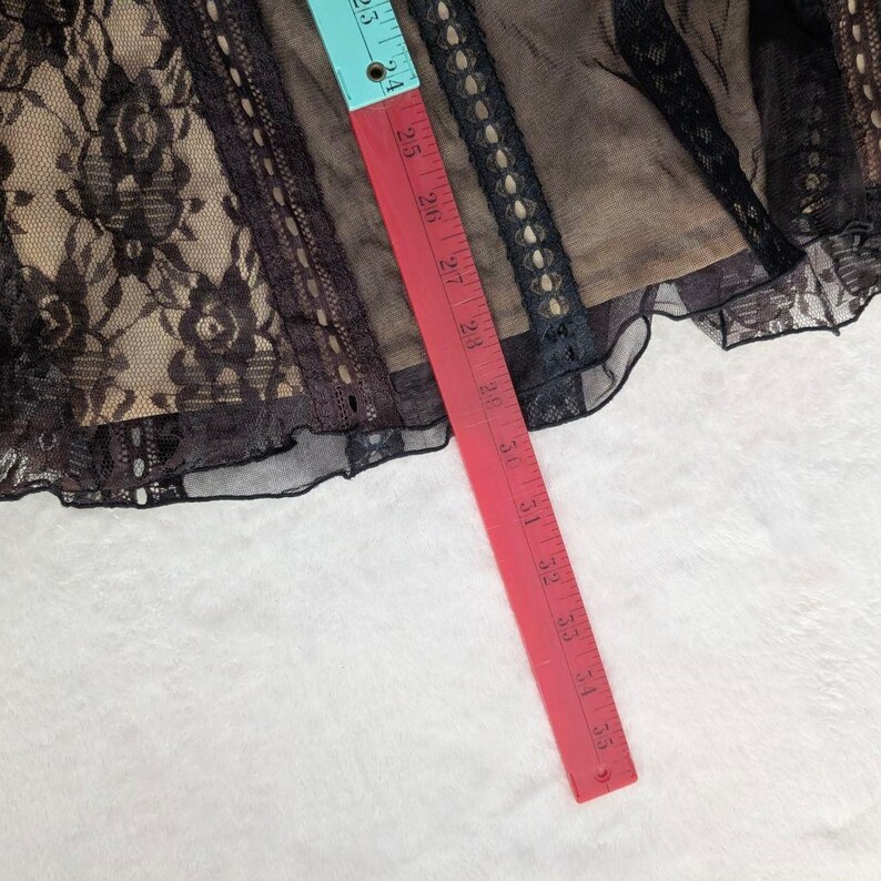 Puede incluir: Tela de encaje negro con una capa base beige, con patrones florales y adornos decorativos. Una cinta m&eacute;trica roja y azul muestra una medida de aproximadamente 61 cm. La tela est&aacute; sobre una superficie blanca.