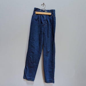 May include: Dark blue denim pants hanging on a wooden hanger. The pants have a high waist and a straight leg design. The denim is a solid, medium wash with visible seams and pockets. The pants are approximately 30 inches long.