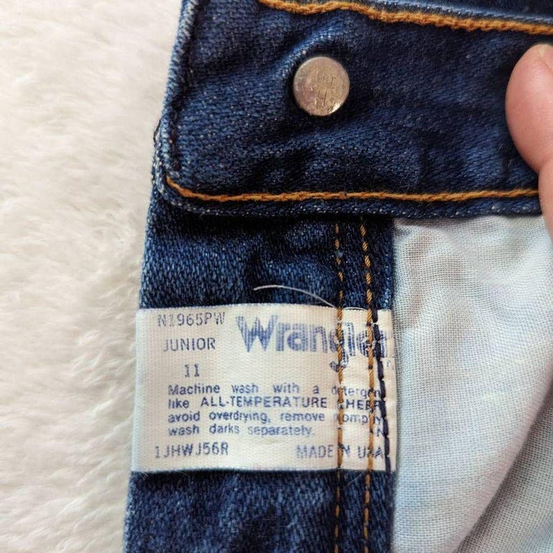 May include: Close-up of a pair of blue denim jeans with a metal button and orange stitching. A white tag reads "Wrangler Junior 11" with washing instructions. Made in the USA.