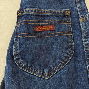 May include: Close-up of a dark blue denim jean pocket with a brown Wrangler label. The pocket has a rounded bottom and is stitched with orange thread. The jeans have a classic design.