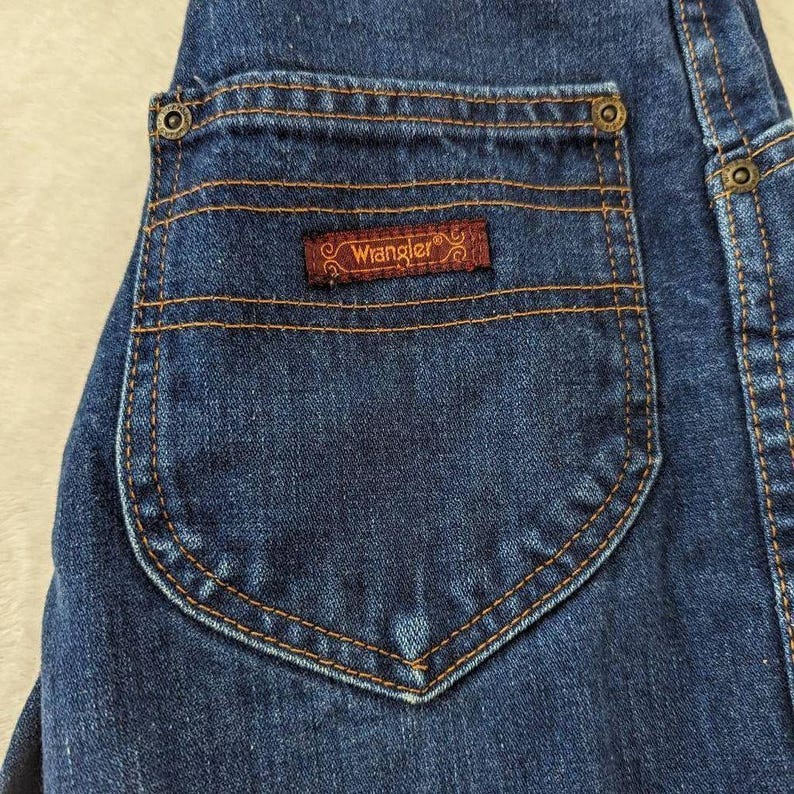 May include: Close-up of a dark blue denim jean pocket with a brown Wrangler label. The pocket has a rounded bottom and is stitched with orange thread. The jeans have a classic design.