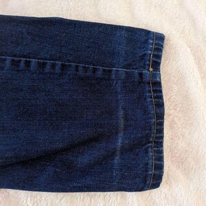 May include: Close-up of a section of dark blue denim fabric. The hem is turned up and stitched with a contrasting yellow thread. The fabric has a textured appearance and is set against a white, fluffy background.
