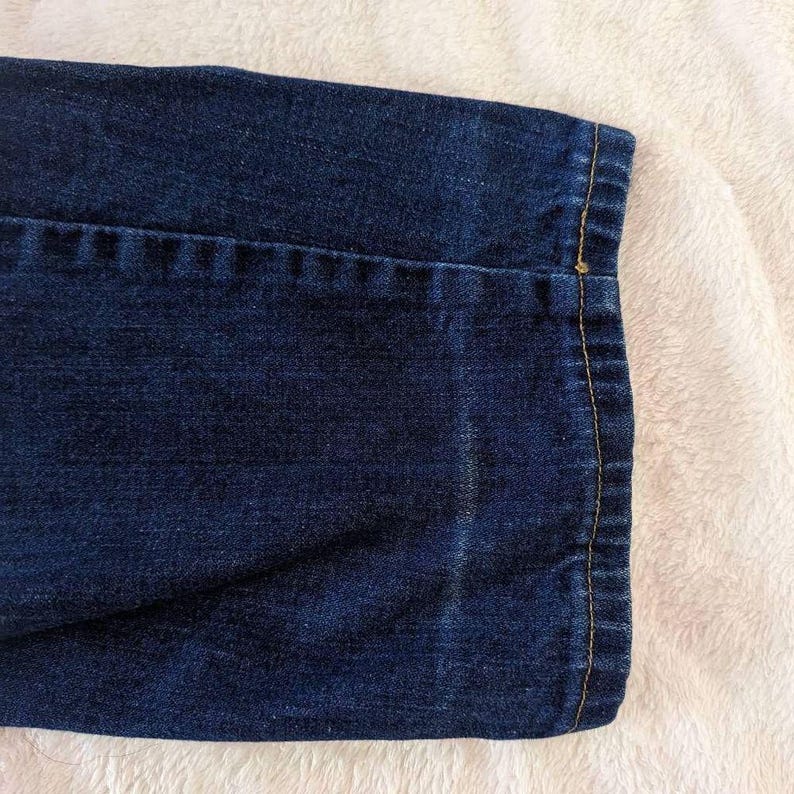 May include: Close-up of a section of dark blue denim fabric. The hem is turned up and stitched with a contrasting yellow thread. The fabric has a textured appearance and is set against a white, fluffy background.