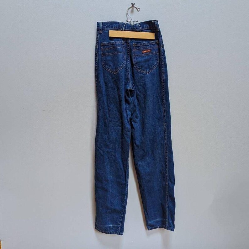 May include: Dark blue denim jeans hanging on a wooden hanger. The jeans have a high waist, two back pockets, and orange stitching. The jeans are displayed against a light gray wall.