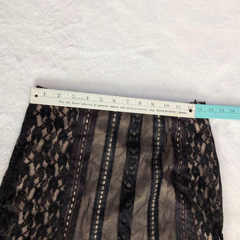 Puede incluir: Falda de encaje negro con forro beige. La falda se mide con una regla, mostrando una longitud de aproximadamente 33 cm. El encaje presenta un patr&oacute;n floral en los lados y rayas verticales en el centro.