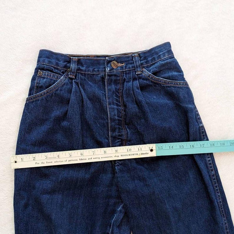 May include: Dark blue denim jeans with a high waist and pleats. A ruler is placed across the jeans, showing measurements in inches. The jeans have pockets and a button closure. The text on the ruler reads "For the finest selection of patterns, fabrics and sewing accessories, shop WOOLWORTH".