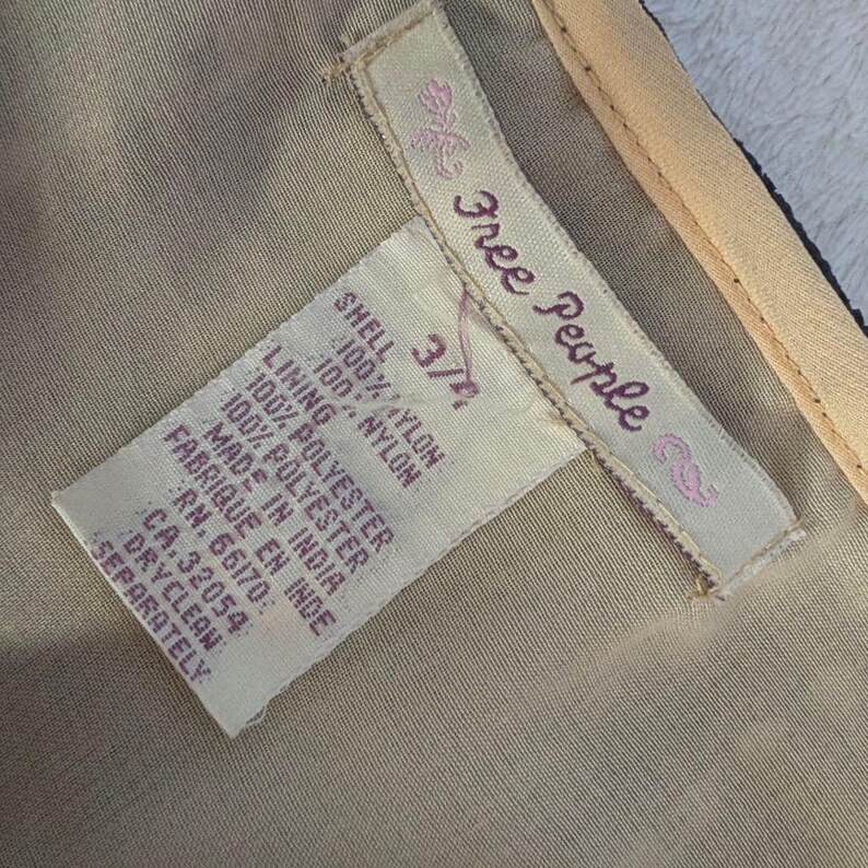 Puede incluir: Primer plano de una etiqueta de ropa de la marca Free People. La etiqueta indica que la carcasa y el forro son de nailon y poli&eacute;ster. Tambi&eacute;n incluye instrucciones de lavado y la talla 3/4.
