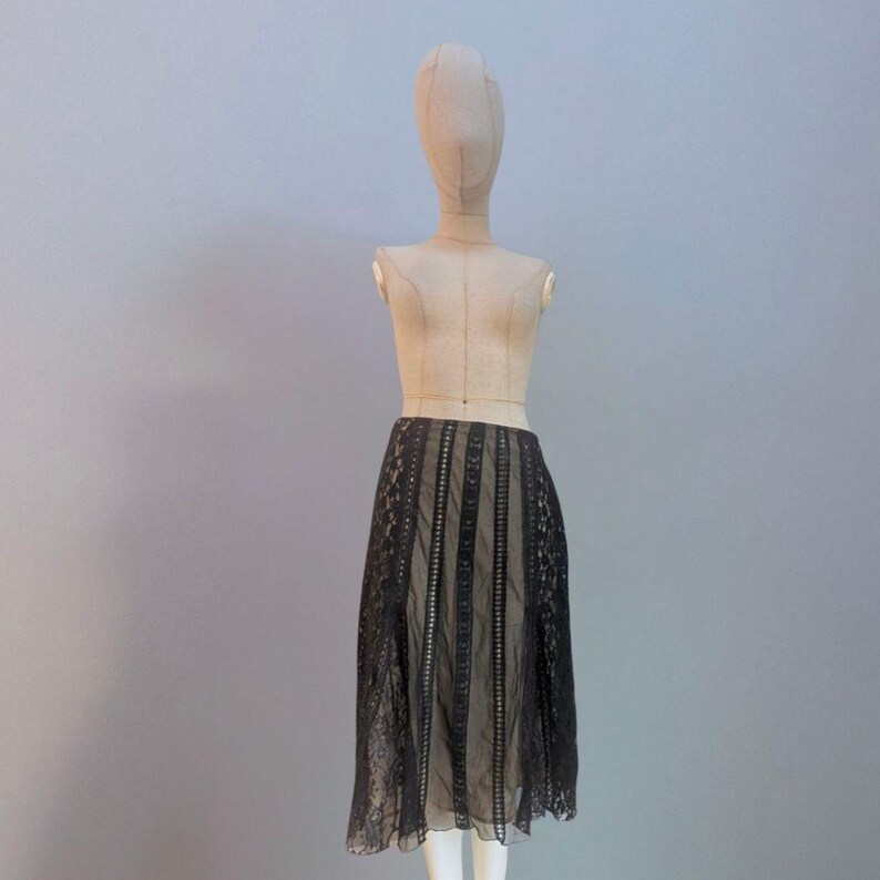 Puede incluir: Falda de encaje negro exhibida en un maniqu&iacute;. La falda presenta una combinaci&oacute;n de paneles lisos y estampados, creando un efecto de capas. La falda es hasta la rodilla y tiene una silueta fluida, mostrando un estilo vintage o bohemio.