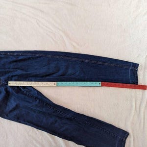 May include: Dark blue denim jeans with a straight leg design. A ruler is placed along the jeans to measure the length. The jeans are laid flat on a white surface.