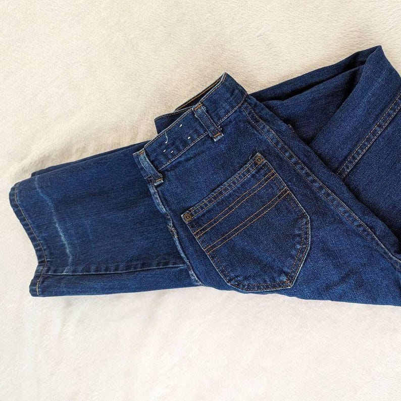May include: Folded pair of dark blue denim jeans. The jeans have a classic five-pocket design with a back pocket and a front pocket. The stitching is a contrasting brown color. The jeans are folded neatly on a white surface.