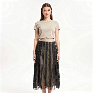 Puede incluir: Una mujer con una camiseta beige de manga corta y una falda midi de encaje negro. La falda tiene paneles verticales de encaje sobre un forro beige. El conjunto se presenta sobre un fondo blanco liso, destacando el dise&ntilde;o y la textura de la ropa.