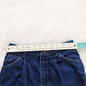 May include: Close-up of a dark blue denim skirt with a measuring tape across the waistband. The skirt has a button closure, pockets, and pleats. The measuring tape reads 12 inches across the top. The text on the tape reads "For the finest selection of patterns, fabrics and sewing accessories, shop WOOLWORTH".