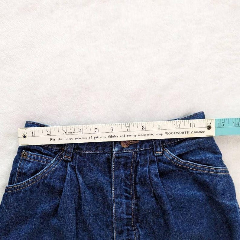 May include: Close-up of a dark blue denim skirt with a measuring tape across the waistband. The skirt has a button closure, pockets, and pleats. The measuring tape reads 12 inches across the top. The text on the tape reads "For the finest selection of patterns, fabrics and sewing accessories, shop WOOLWORTH".